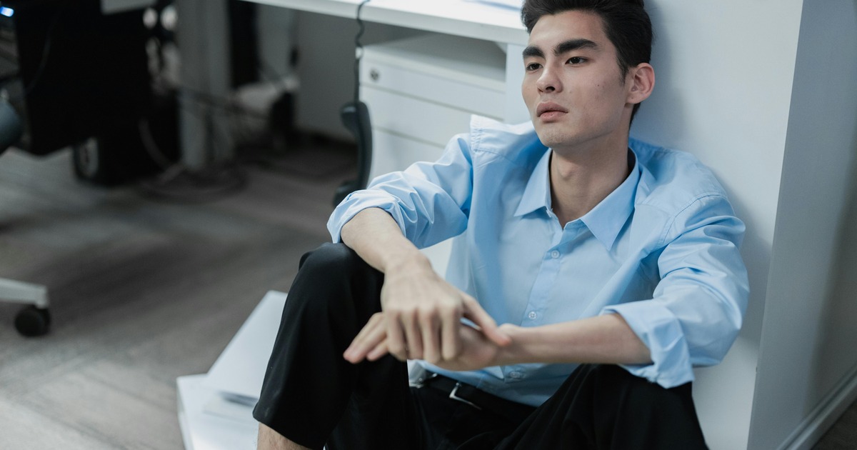 An exhausted Asian man sits on an office floor looking reflective and stressed, surrounded by paperwork.